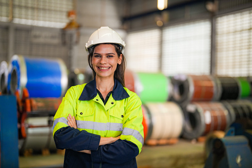 engineer woman standing with confidence with green working suite dress and safety helmet in front of warehouse of steel role material. smart industry worker operating.