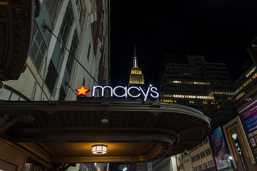 New York, Manhattan, USA. 05.07.2023. Night view of exterior of Macy's sign on store wall with Empire State Building in background between skyscrapers.