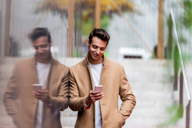 istock Portrait of stylish handsome young man with coat standing outdoors and leaning on wall using a smartphone 1488405055