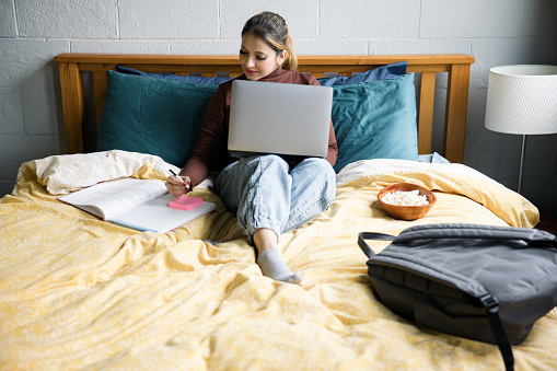 An adult Indian woman does research for school utilizing the internet on her computer and her textbooks.  She lays comfortably on her bed with snacks, enjoying a leisurely study session.