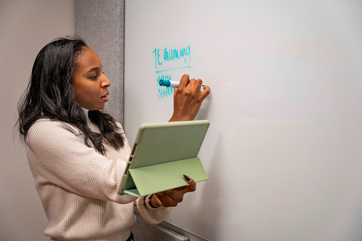 African American woman writing on whiteboard holding tablet