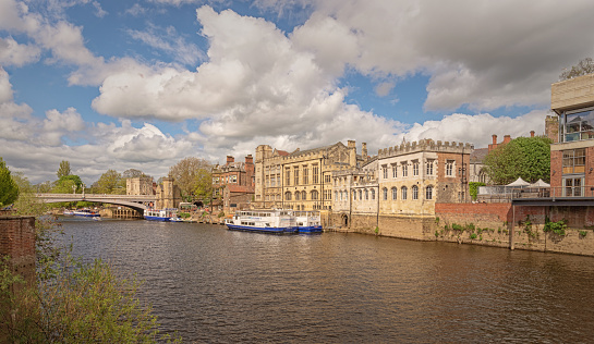The Guildhall in York with Lendal Bridge in the background.  Tourist boats are moored by a wharf and a shrub is in the foreground. A sky with cloud is above.
