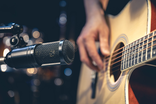 Male musician playing acoustic guitar behind microphone, close-up, recording in a music studio.