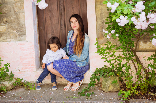 Attractive woman and little handsome baby boy sitting near the ancient door with white roses in the old town on a walk outdoor
