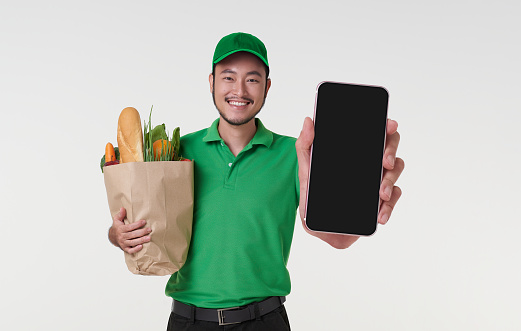 Asian delivery man showing smartphone application and holding fresh food paper bag isolated on white background. express shopping online delivery concept.