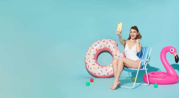 femme heureuse se relaxant dans une chaise longue près de l’anneau en caoutchouc prenant une photo selfie en regardant un smartphone sur un fond d’espace de copie bleu. femme se relaxant sur la plage en vacances d’été. - inflatable ring photos photos et images de collection