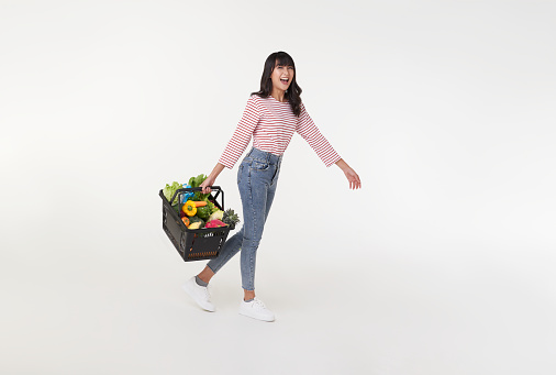 Happy Asian woman holding shopping basket full of vegetables and groceries walking at supermarket isolated on white studio background. copy space