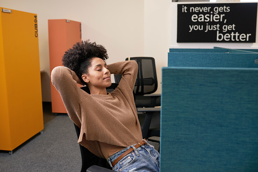 Young multi-ethnic woman leans with arms behind head, taking break in office. High quality photo