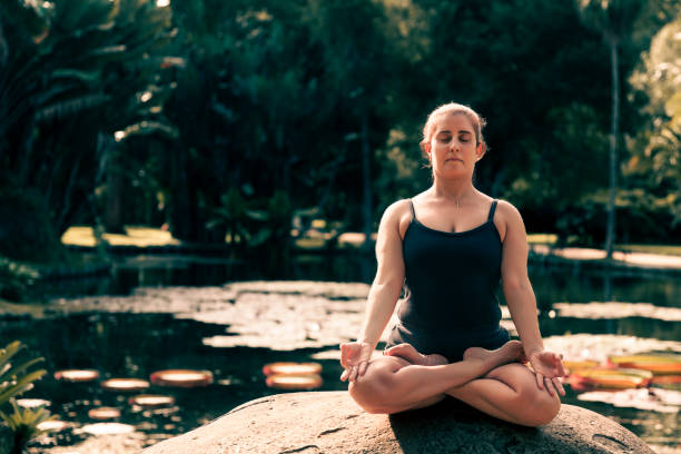 mulher de meia-idade meditando sentada em uma rocha à beira de um lago - lake tranquil scene landscape zen like - fotografias e filmes do acervo