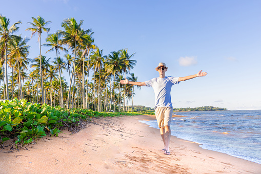 Man with open arms walking on a beautiful beach and enjoying the moment, with coconut trees in the background. Enjoying on the beach.