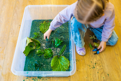 Little girl playing in handmade swamp of green-dyed chia seeds with insect, fish and plant models. Sensory development and experiences, themed activities with children, fine motor skills development.