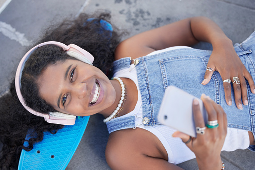 Portrait, phone and music with a black woman skater lying on her board at a skatepark from above. Face, skating and social media with a female skateboarder resting on the ground during a break