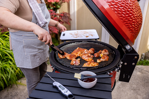 High quality stock photo of a Asian American woman cooking chicken outdoors on a charcoal grill demonstrating food safety by using a plastic cutting board and keeping the area clean.
