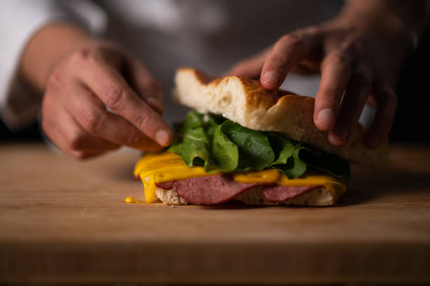 A male chef preparing a sandwich with focaccia bread, rose beef and cheddar cheese on a dark background.