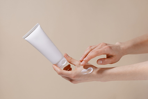 Las manos frágiles de una mujer sostienen un tubo blanco de crema facial y aplican crema hidratante a su piel sobre un fondo beige aislado. Imagen para tu diseño. photo