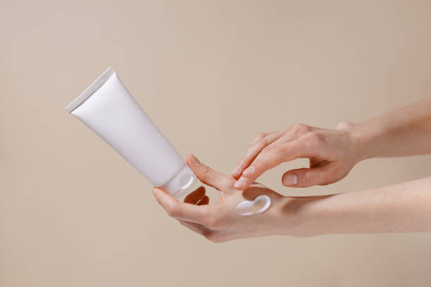 fragile hands of a woman hold a white mockup tube of facial cream and apply moisturizer to her skin on a beige isolated background. image for your design. - 潤手霜 個照片及圖片檔