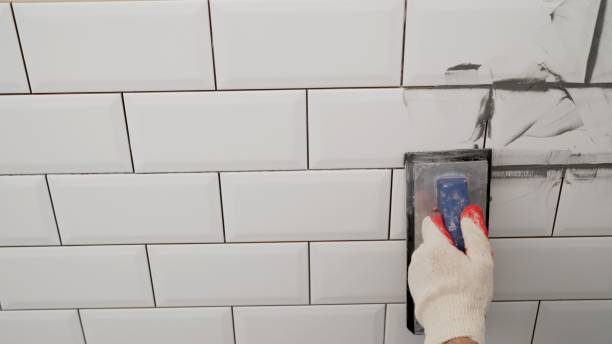 arbeiterhände mit einem gummispatel und verfugen mit paste zwischen weißen keramikfliesen. nahaufnahme von fugenmörtel. verfugungsnähte auf einer fliesenrückwand. - grouting stock-fotos und bilder