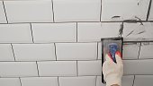 Workers' hands using a rubber spatula and grouting with paste between white ceramic tiles. Close-up of grout. Grouting seams on a tile backsplash.