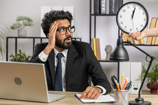 Indian businessman working on office laptop with anxiety checking time on clock, running late to work, being in delay, deadline. Manager freelancer man looking at hour, minutes worrying to be punctual