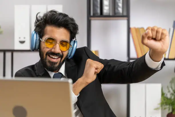 Photo of Indian businessman wearing headphones listening music dancing after working on laptop at home office