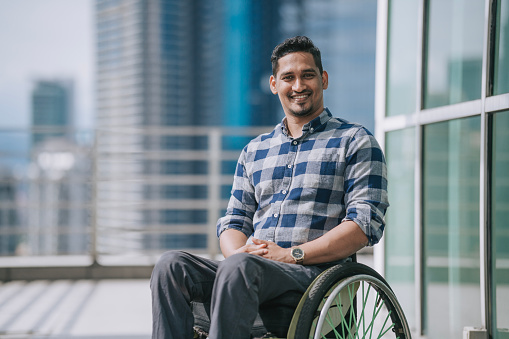 Portrait Asian Indian white collar worker in wheelchair looking at camera smiling with cityscape background