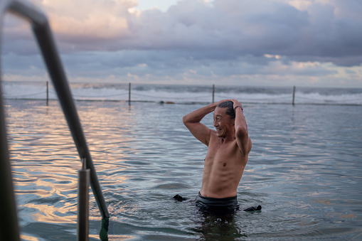 A male of Japanese ethnicity takes an evening swim on the eastern beaches of Australia. He is smiling and standing in a rock pool that is located in front of the ocean. He has his wetsuit pulled down and he is tying up his hair.