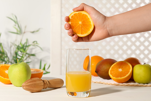 Woman squeezing orange over glass of juice at white wooden table, closeup