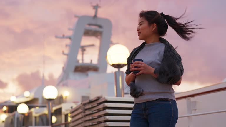 Peaceful serene Asian woman traveller hand hold hot drink enjoy watching sunset on cruise deck ship cheerful happiness moment