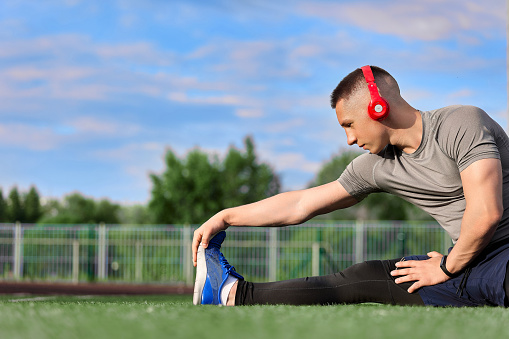 Handsome young Caucasian man in sportswear and headphones stretches while warming up outdoors. A healthy man is engaged in fitness, stretching in the park against the blue sky