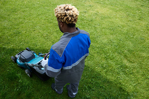 Professional lawn care service. Top view of an African man in overalls mowing green grass in a modern garden with a lawn mower. A black man with an Afro hairstyle uses a lawn mower in the backyard.
