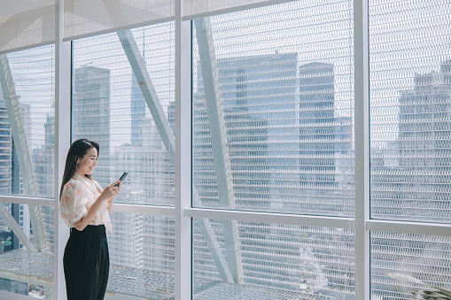 Asian Chinese business woman standing in front of window in office phone messaging