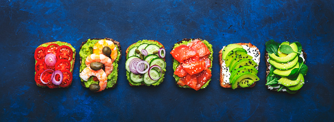 Avocado toast set with salmon, shrimps, tomatoes, cucumbers, soft cheese and spinach, cashew and sesame seeds, blue table background, top view