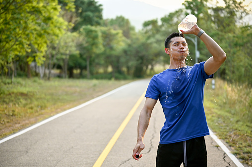 Tired and exhausted millennial Asian male runner pouring water from a bottle on his face, refreshing himself with water while running in a park.
