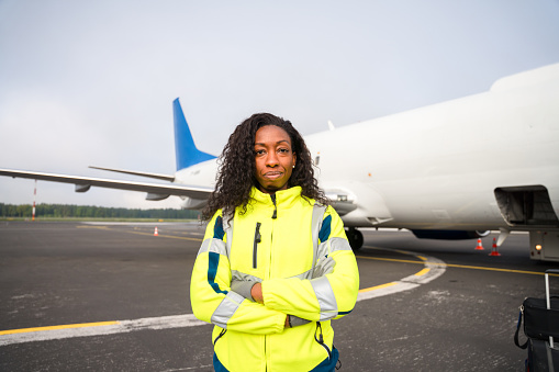 African American aircraft maintenance technicians standing standing next to an airplane at the airport runway wearing a reflective vest with hands crossed looking at camera.