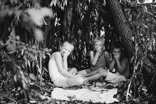 Three children play in a hut which they themselves have built from leaves and twigs. Black and white photography