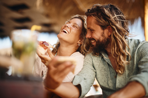 Cheerful couple having fun while eating pizza in summer day on the beach.
