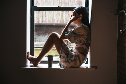 Sad woman sitting by the window in her apartment.