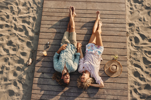 High angle view of young happy couple holding hands while lying in summer day on the beach. Copy space.