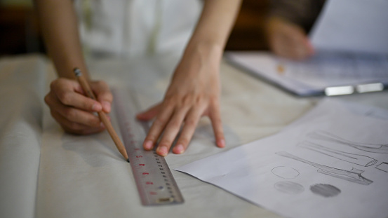 Close-up image of a female fashion designer or tailor using pencil and ruler, measuring and making paper pattern, working in her studio.