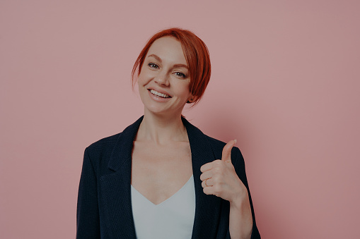 Young happy friendly ginger woman showing thumb up and looking at camera with smile, recommending something with gesture, isolated on pink background, cheerful female being satisfied with choice