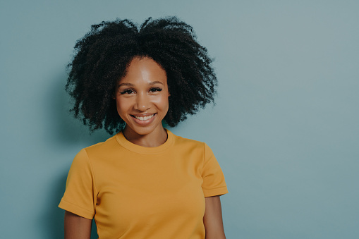 Happy young dark skinned woman with curly hairstyle smiling at camera with pleased face expression, african female in yellow shirt expressing happiness, posing against blue studio background