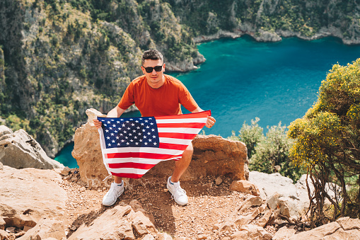 Young man sitting on a mountain top and holding the US flag in his hands. Veteran traveller holding the American flag while sitting on a mountain top. 4 fourth of July Independence Day.