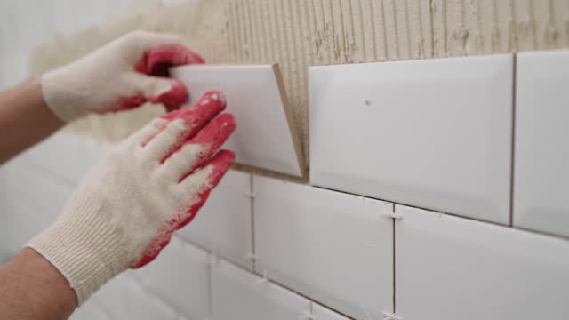 Gluing ceramic tiles to the wall in the kitchen. The hands of a handyman smear tile glue with a spatula on the wall next to the glued tile. Construction details, repair work.