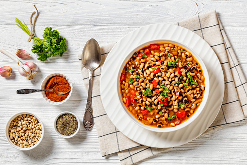 Vegan Hoppin John, savory and spicy black-eyed pea stew in white bowl on white wood table with ingredients and spices, southern America recipe, horizontal view from above, flat lay