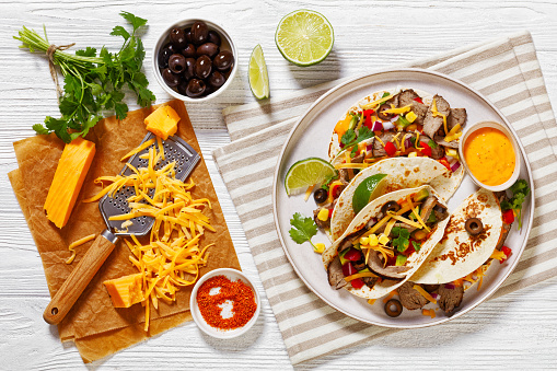 Grilled Steak Tacos with olives, tomatoes, red onion, avocado, cilantro, corn, shredded cheddar cheese and thousand island sauce on platter on white wood table with ingredients, flat lay