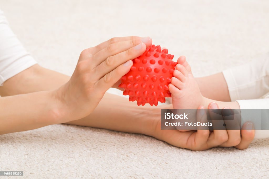 Masseur hand holding red rubber massage ball and massaging infant foot on carpet. Baby healthcare. Closeup. Side view. Massage Ball Stock Photo