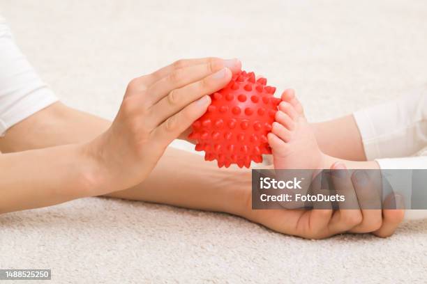 Masseur Hand Holding Red Rubber Massage Ball And Massaging Infant Foot On Carpet Baby Healthcare Closeup Side View Stock Photo - Download Image Now