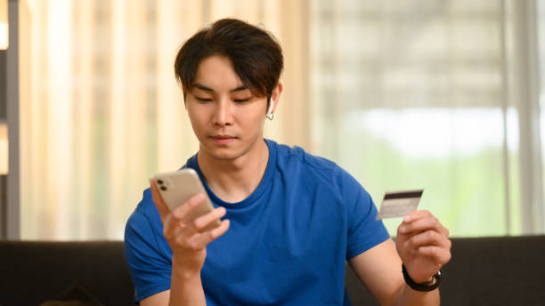 istock Handsome smiling man holding credit card and using mobile phone, making online transactions, shopping online at home. 1488525421