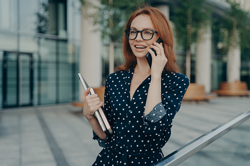 Horizontal shot of cheerful redhead European woman talks on modern mobile phone has glad expression smiles broadly holds laptop and diary walks outside against blurred background during daytime