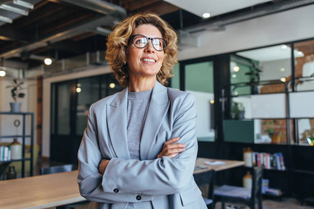 Business woman in her 40's looks away thoughtfully in an office Business woman in her 40's looks away thoughtfully in an office. Professional woman standing in a suit with crossed arms. businesswoman stock pictures, royalty-free photos & images
